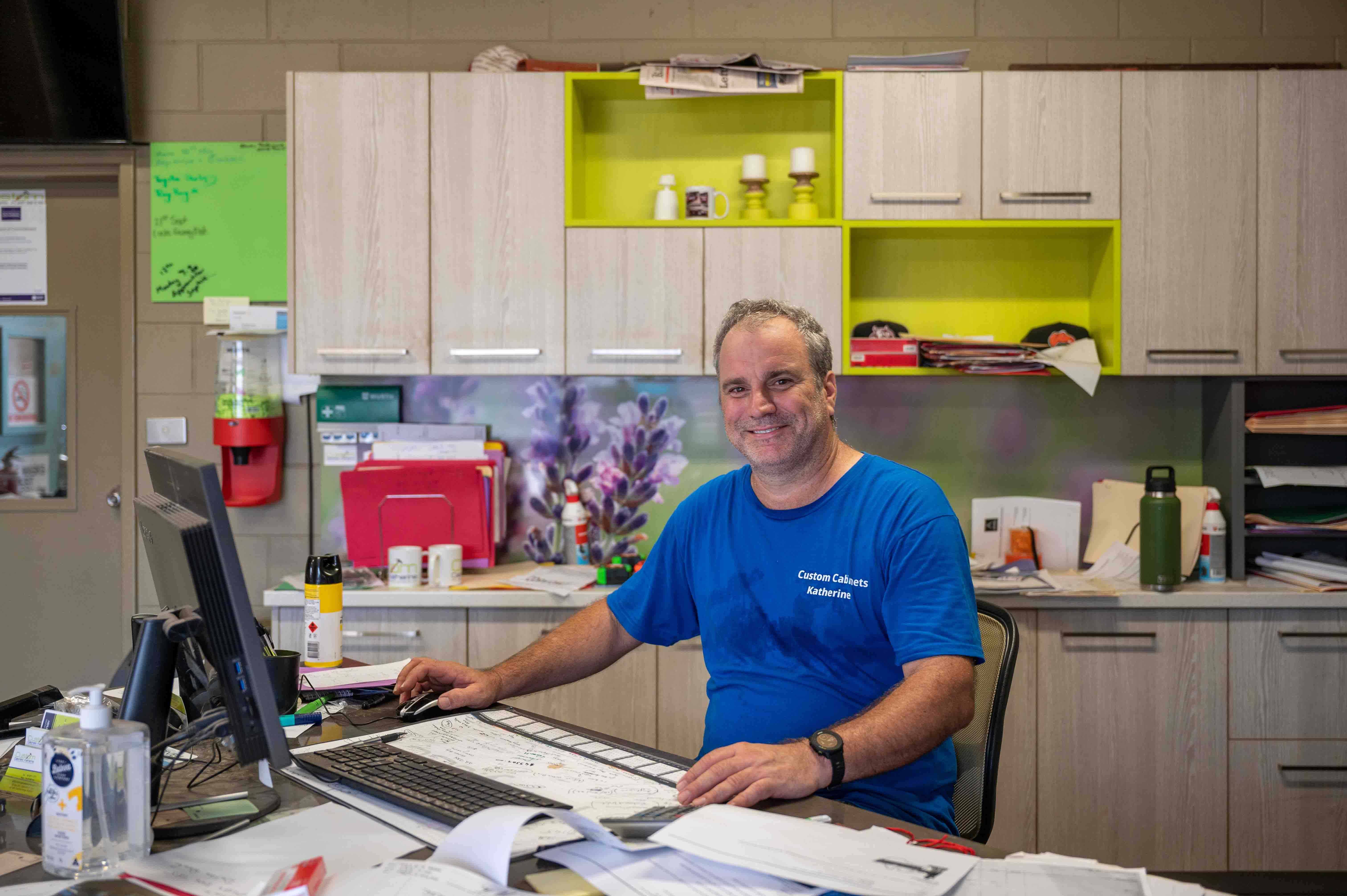  Man in a blue shire sits at a desk surrounded by papers.